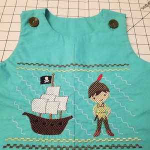 May include: A turquoise sleeveless onesie with an embroidered design of a pirate ship and a boy holding a sword. The ship has a black flag with a skull and crossbones. The boy is wearing a brown hat with a feather.