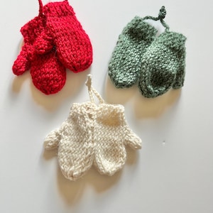 May include: Three pairs of knitted mittens in red, green, and off-white. Each pair has a loop for hanging. The mittens are made of a chunky knit yarn, and are likely handmade. The mittens are small, possibly for decoration.
