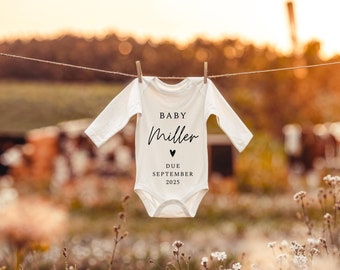 May include: A white baby onesie hanging on a clothesline in a field of wildflowers. The onesie has the text "Baby Miller" and "Due September 2023" printed on it.