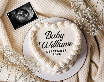 May include: A white heart-shaped cake with white frosting and the words "Baby Williams September 2025" written in black on the front. The cake is on a white plate and a white blanket.