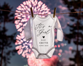 Puede incluir: Un body blanco para bebé con el texto "It's a Girl" en letras cursivas negras, un pequeño corazón negro y el texto "Baby Smith June 2025" en letras negras. El body está colgado en un tendedero contra un fondo borroso de fuegos artificiales rosas.