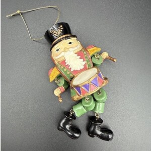 May include: A nutcracker ornament featuring a black hat with gold detail, a white beard, and a red and green uniform. The figure holds a drum with purple and red triangles and wears green trousers with black boots, suspended by a gold string.