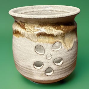 May include: A handmade ceramic pot with a wide opening and a textured surface. The pot features a light beige base with brown and tan accents around the top. It has several circular cutouts on the front, creating a decorative design.