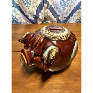 Pode incluir: Um cofrinho de cerâmica marrom com uma ranhura para moedas na parte superior e um anel de metal no focinho. O banco tem um acabamento brilhante com detalhes brancos e bege ao redor da parte superior e dos olhos. O cofrinho está em uma superfície de madeira.