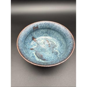 Könnte beinhalten: Eine handgefertigte Keramikschale mit glänzender, blauer Glasur. Die Schale hat eine abgerundete Form mit einem braunen Rand und Innenakzenten. Der Innenraum weist ein gesprenkeltes Muster aus Blau- und Brauntönen auf, das einen einzigartigen, künstlerischen Look erzeugt.