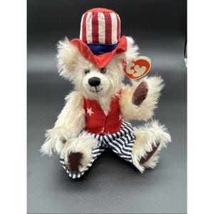 Könnte beinhalten: Ein Plüschteddybär in patriotischer Kleidung. Der Bär hat cremefarbenes Fell, eine rote Weste mit weißen Sternen und eine blau-weiß gestreifte Hose. Er trägt einen rot-weiß-blauen Uncle-Sam-Hut.