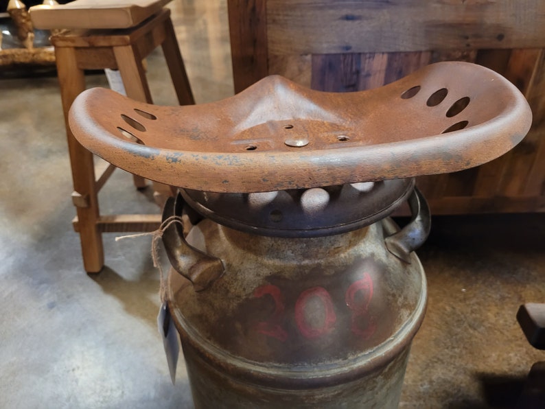 Puede incluir: Un asiento de tractor de metal oxidado unido a una lata de leche reutilizada. La lata tiene el n&uacute;mero rojo "202" pintado en ella.