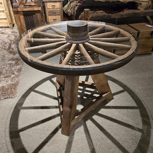 May include: A rustic wooden wagon wheel table with a glass top. The table features a dark brown rim and spokes, supported by a triangular wooden base. The table's diameter is approximately 48 inches.