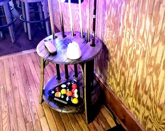 May include: A rustic, wooden pool cue rack with a round top and a lower shelf. The rack holds several pool cues and a triangle of billiard balls. A small, white object and a wooden item sit on the top shelf.
