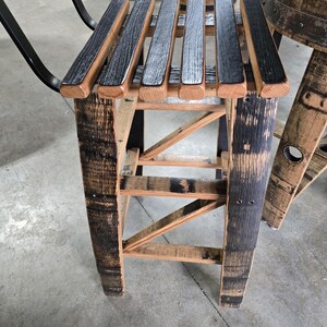 May include: A rustic wooden stool made from reclaimed barrel staves. The seat is composed of alternating black and natural wood slats. The legs and frame feature a natural wood finish with dark charred accents, showcasing the barrel's original markings.