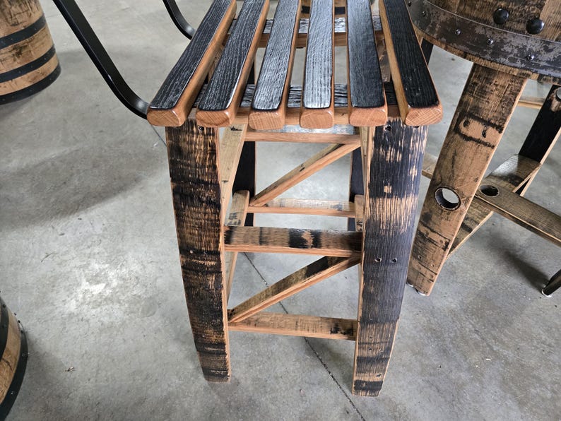May include: A rustic wooden stool made from reclaimed barrel staves. The seat is composed of alternating black and natural wood slats. The legs and frame feature a natural wood finish with dark charred accents, showcasing the barrel's original markings.