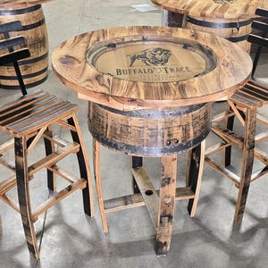 May include: A rustic, round bar table and two matching stools made from reclaimed wood. The table features a clear inset with the "Buffalo Trace" logo. The stools have slatted seats and black accents. The table is approximately 36 inches in diameter.