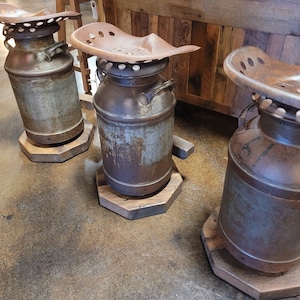 Puede incluir: Tres taburetes r&uacute;sticos de metal en forma de bid&oacute;n de leche con bases de madera. Cada taburete tiene un asiento de tractor de metal oxidado.
