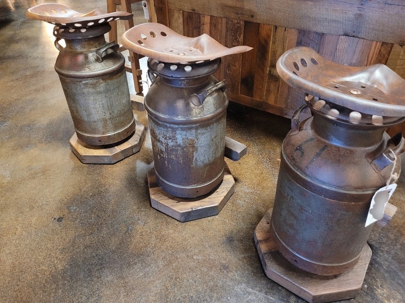 Puede incluir: Tres taburetes r&uacute;sticos de metal en forma de bid&oacute;n de leche con bases de madera. Cada taburete tiene un asiento de tractor de metal oxidado.