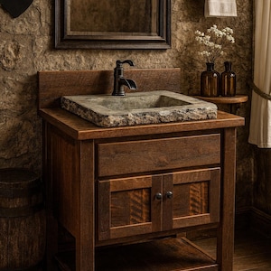 May include: A wooden bathroom vanity cabinet with a dark brown finish, featuring a rectangular stone vessel sink with a rough-hewn edge and a dark metal faucet. The vanity includes a top drawer, two lower cabinet doors with small knobs, and an open shelf.
