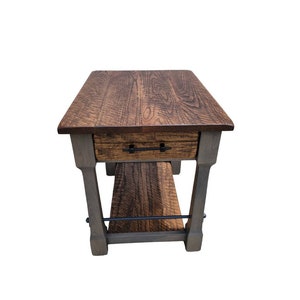 May include: A rustic wooden side table with a drawer and lower shelf. The tabletop and drawer front feature a natural wood grain finish. The legs are painted gray, and a black metal bar connects the legs.