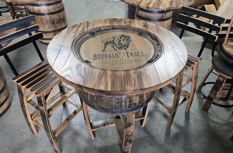 May include: A round wooden table with a glass top, featuring the "Buffalo Trace" logo. The table is supported by a repurposed barrel base and is surrounded by matching wooden chairs. The table is approximately 36 inches in diameter.