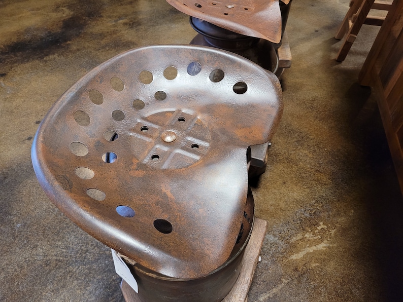Puede incluir: Un asiento de tractor de metal oxidado con un dise&ntilde;o circular y m&uacute;ltiples agujeros. El asiento est&aacute; montado sobre una base de metal.