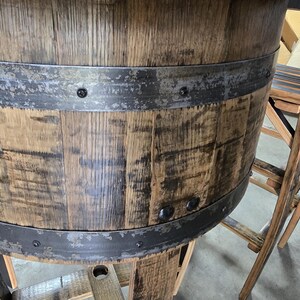 May include: A rustic wooden barrel table with metal bands. The table has a weathered appearance, showcasing the natural grain and texture of the wood. A wooden stool is next to the table. The design suggests a bar or pub setting.
