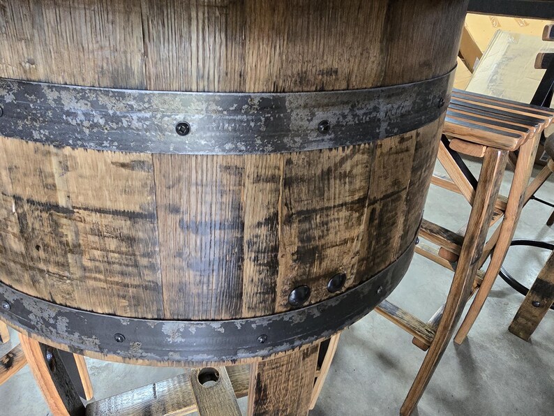 May include: A rustic wooden barrel table with metal bands. The table has a weathered appearance, showcasing the natural grain and texture of the wood. A wooden stool is next to the table. The design suggests a bar or pub setting.