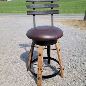 May include: A rustic bar stool with a dark brown leather seat and a backrest made of three horizontal wooden slats. The legs are crafted from light-colored wood and are connected by a black metal ring. The stool is approximately 30 inches tall.