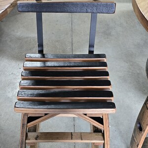 May include: A wooden chair with a slatted seat and back, constructed from repurposed barrel staves. The chair features a dark, charred finish on the slats and a natural wood tone on the frame, with metal supports. The chair is designed for indoor or outdoor use.