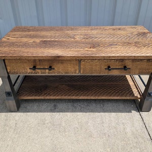 May include: A rustic coffee table with a wood top and metal legs. The table has two drawers and a lower shelf. The wood is a dark brown color and the metal is a dark gray color.