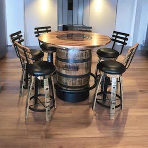 May include: A round wooden table with a glass top sits on a repurposed whiskey barrel base. Four black metal bar stools with brown leather seats surround the table. The stools are also made with repurposed whiskey barrels.