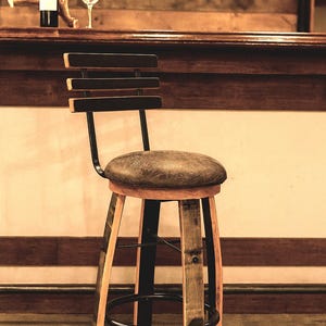 May include: A rustic bar stool with a round, leather-look seat and a backrest with three horizontal slats. The stool features a combination of light wood and dark metal, with a footrest and barrel stave legs. A wine bottle and glass are on the bar.