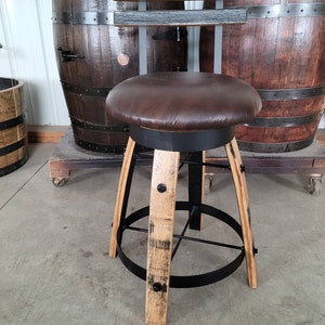 May include: A rustic bar stool with a brown leather seat and a black metal frame. The legs are made from reclaimed wood, and the stool has a footrest. The background features a wooden barrel.