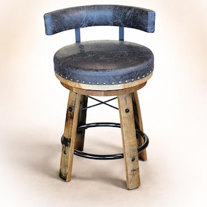 May include: A rustic bar stool with a dark gray leather seat and backrest. The seat is round and trimmed with brass-colored studs. The base is made of weathered wood with a black metal footrest and supports. The stool has a height of approximately 30 inches.