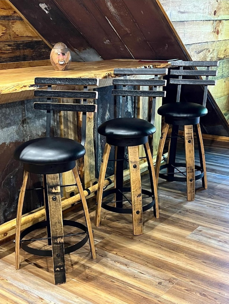 May include: Three rustic bar stools with black leather seats and backs. The stools have wooden legs with metal accents and a footrest. The backs are made of horizontal wooden slats. The stools are set against a wooden bar.