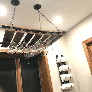 May include: A rustic wine glass rack made from reclaimed wood, suspended from the ceiling by chains. The rack holds multiple wine glasses, showcasing a home bar or kitchen decor. The wood has a natural, weathered appearance.