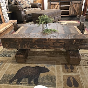 May include: Rustic wooden coffee table with a distressed finish. The table features a rectangular top made of reclaimed wood planks and is supported by thick, block-style legs. A rug with a bear design is beneath the table.