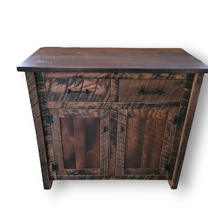 May include: A rustic wooden cabinet with two drawers and two doors. The cabinet is made of reclaimed wood and has a dark brown finish. The doors have black metal hinges and the drawers have black metal pulls.