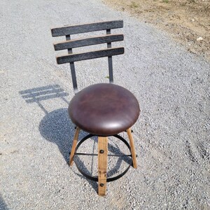 May include: A rustic bar stool with a dark brown leather seat and a backrest made of horizontal wooden slats. The legs are constructed from light-colored wood and feature a metal footrest. The stool is designed for indoor or outdoor use.
