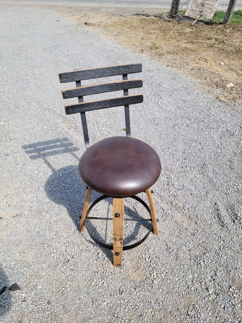 May include: A rustic bar stool with a dark brown leather seat and a backrest made of horizontal wooden slats. The legs are constructed from light-colored wood and feature a metal footrest. The stool is designed for indoor or outdoor use.