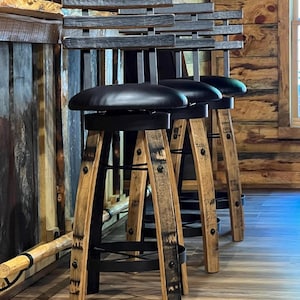May include: Three rustic bar stools with black leather seats and wooden barrel-style legs. The backs are made of horizontal wooden slats. The stools are set against a wooden bar and wall, creating a warm, inviting atmosphere.