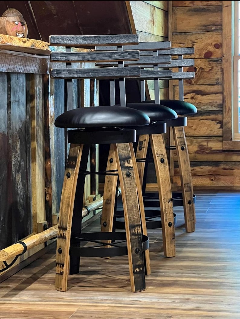 May include: Three rustic bar stools with black leather seats and wooden barrel-style legs. The backs are made of horizontal wooden slats. The stools are set against a wooden bar and wall, creating a warm, inviting atmosphere.