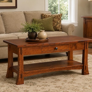May include: A rectangular, wooden coffee table with a drawer and lower shelf. The table is a warm brown color and has a traditional design. A small plant and vase sit on top of the table. The table is in a living room setting.