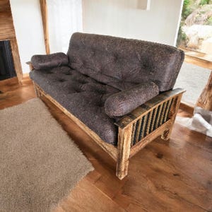 May include: A brown futon sofa with a tufted seat and two bolster pillows. The wooden frame has a rustic design with vertical slats on the arms. The sofa sits on a hardwood floor.