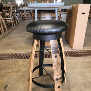May include: A rustic bar stool with a black leather seat and a backrest made of gray-stained wood slats. The stool's base is crafted from repurposed barrel staves, accented with black metal supports and hardware. The stool is approximately 30 inches tall.
