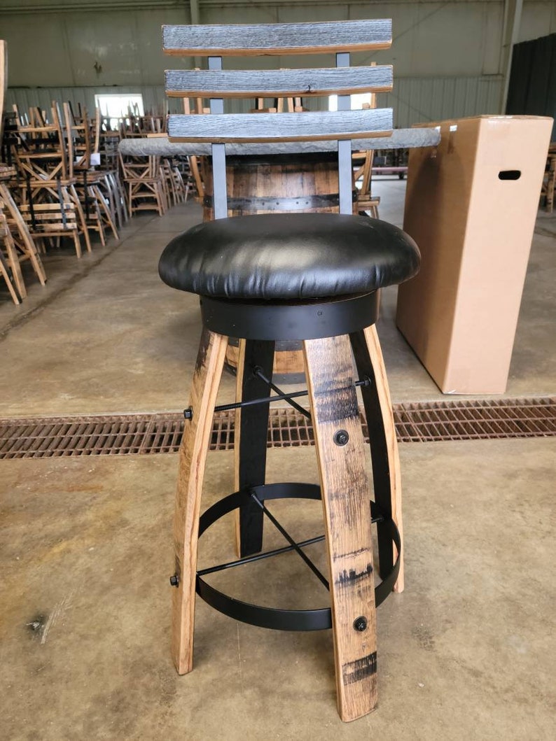 May include: A rustic bar stool with a black leather seat and a backrest made of gray-stained wood slats. The stool's base is crafted from repurposed barrel staves, accented with black metal supports and hardware. The stool is approximately 30 inches tall.