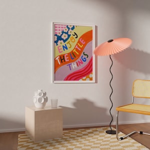 以下が含まれることがあります： 白いフレームに「Enjoy the little things」という文字が書かれたカラフルな抽象的なアートプリント。プリントは、ピンクのランプ、白い花瓶、籐の椅子のある白い壁にかけられています。