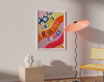 May include: A colorful abstract art print with the text "Enjoy the little things" in a white frame. The print is hanging on a white wall with a pink lamp, a white vase, and a chair with a wicker seat.