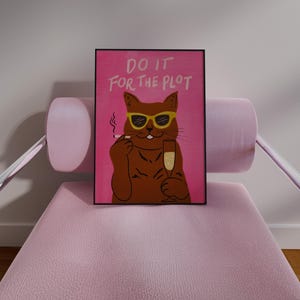 May include: A framed art print featuring a brown cat wearing sunglasses, holding a champagne flute and a cigarette. The pink background has the text "DO IT FOR THE PLOT" in white. The artwork is displayed on a pink chair.