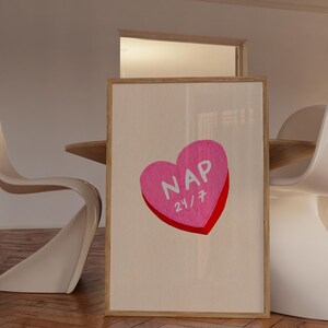 Könnte beinhalten: Ein gerahmter Druck mit einem rosa herzförmigen Bonbon-Design. Das Bonbon hat das Wort "NAP" und "24/7" in weißer Schrift mit roter Umrandung. Das Kunstwerk wird in einem Raum mit weißen Stühlen und einem Holztisch ausgestellt.
