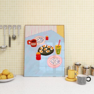 Op de afbeelding: Een ingelijste illustratie van een keukentafelsetting met een rode mok, een bord met eieren, een glas sap en een bord met blauwe bessen. De illustratie is op een lichtblauwe achtergrond met een wit en roze geruit patroon.