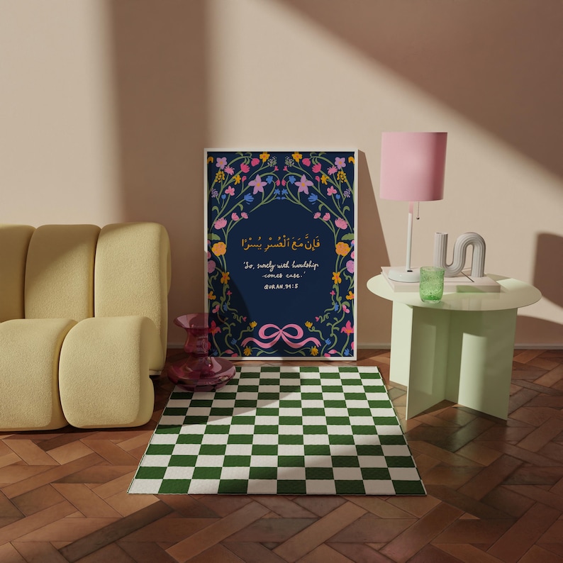 May include: A framed print with floral border and Arabic text, "So, surely with hardship comes ease." The print is on a dark blue background. A green and white checkered rug is in front of the print.