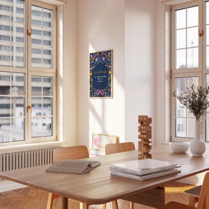 May include: A framed art print with Arabic calligraphy and floral accents hangs on a white wall. The print features a dark blue background with colorful flowers and text. The room has a wooden table, chairs, and large windows.